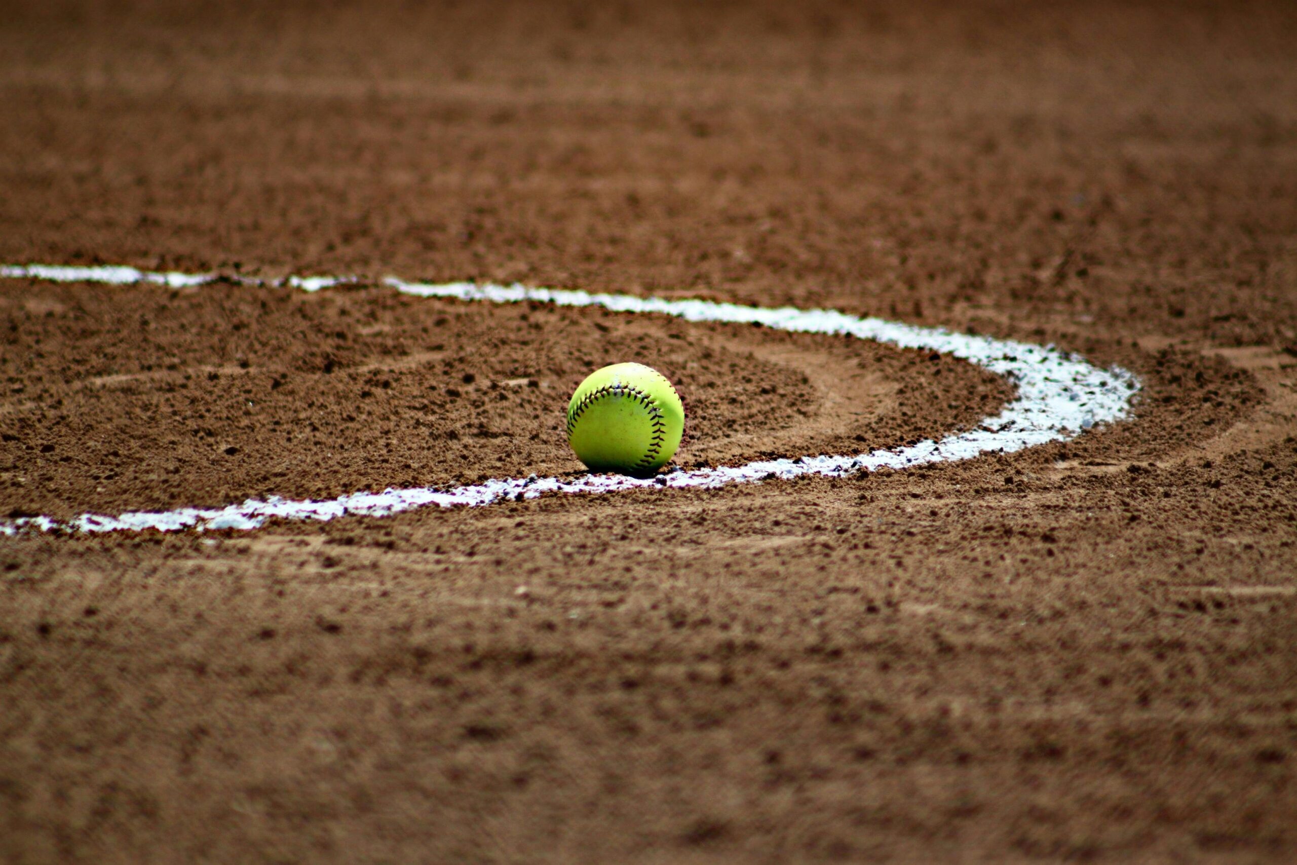 A single lime green colored softball rests on a dirt field. A white curved line is visible in the brown dirt that extends from the left side of the image.